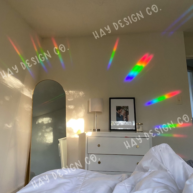 Puede incluir: Un dormitorio con una c&oacute;moda blanca, un espejo dorado y una cama blanca con ropa de cama blanca. Las paredes son blancas y hay reflejos de prisma arco&iacute;ris en las paredes. El texto "HAY DESIGN CO." est&aacute; escrito en las paredes.