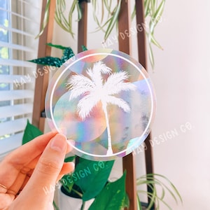 Puede incluir: Una pegatina redonda iridiscente con una silueta de palmera blanca en el centro. La pegatina tiene un borde blanco y el texto "HAY DESIGN CO." está impreso alrededor del borde.