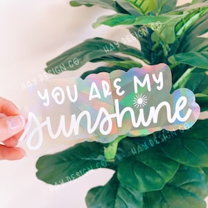 以下が含まれることがあります： 虹色の虹色の背景と「You are my sunshine」という白いテキストと小さな太陽のアイコンが書かれたホログラフィックステッカーです。
