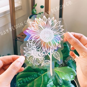 May include: A holographic sticker featuring a white line drawing of two sunflowers. The sticker has a rainbow iridescent background and the text "HAY DESIGN CO." is printed on the sticker.