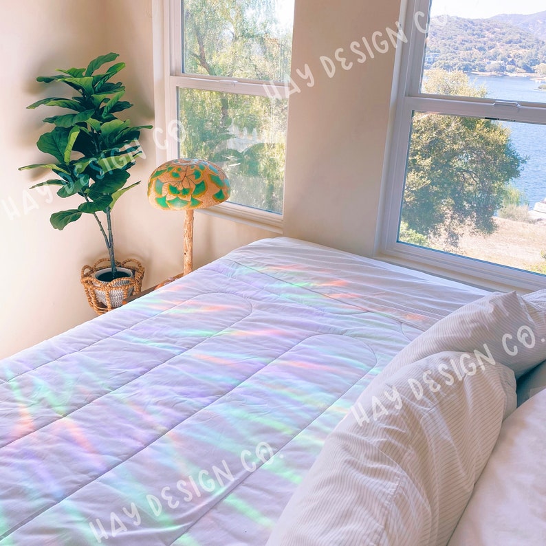 Puede incluir: Una cama blanca con un edred&oacute;n blanco y una almohada blanca. El edred&oacute;n tiene un patr&oacute;n de arco&iacute;ris. Hay una l&aacute;mpara de hongo verde y marr&oacute;n en una mesita de noche junto a la cama. Una ventana con vista a un lago y monta&ntilde;as est&aacute; detr&aacute;s de la cama.
