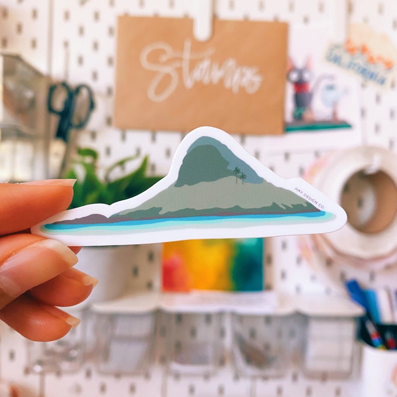 May include: A white sticker with a green and blue illustration of a small island with a mountain and palm trees. The sticker has the text "MAY DESIGN CO." in the bottom right corner.