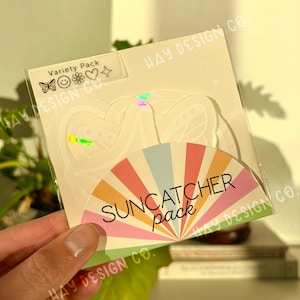Könnte beinhalten: Eine transparente Kunststoffverpackung mit einem Sonnenfänger-Pack. Die Verpackung hat ein Etikett "Variety Pack" mit Schmetterling-, Smiley-, Blumen-, Herz- und Sternsymbolen. Das Sonnenfänger-Pack hat ein Regenbogenfächer-Design mit den Worten "SUNCATCHER pack".