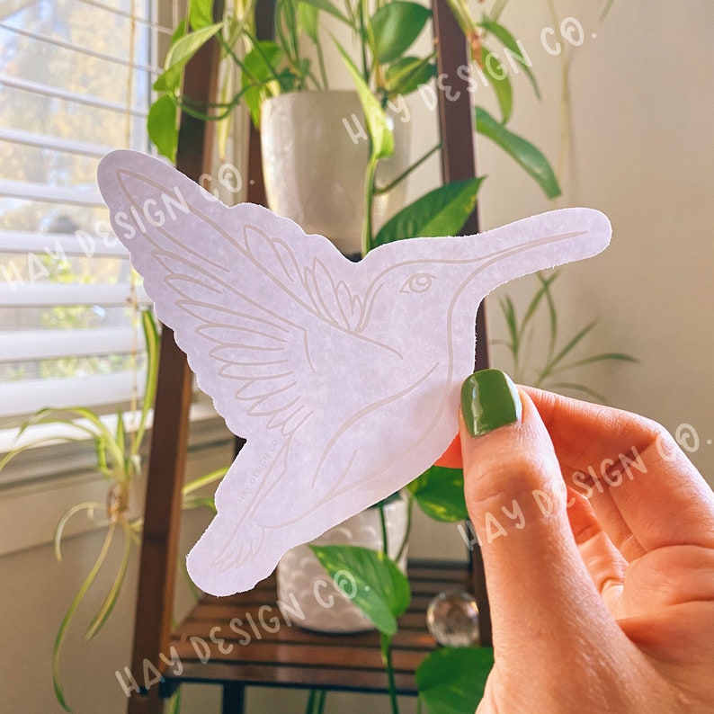 Puede incluir: Pegatina de colibr&iacute; blanca con un contorno detallado de las alas y el cuerpo del p&aacute;jaro. La pegatina est&aacute; sobre un fondo blanco y la est&aacute; sujetando una mano.