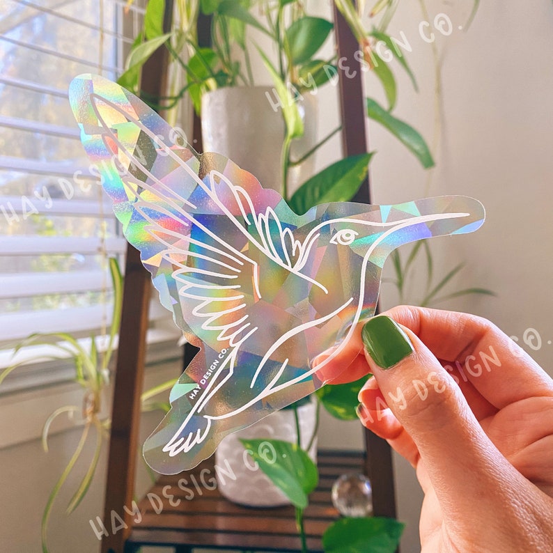 Puede incluir: Una pegatina hologr&aacute;fica con un contorno blanco de un colibr&iacute; en vuelo. La pegatina tiene un acabado iridiscente arco&iacute;ris y el texto "HAY DESIGN CO." est&aacute; impreso en la pegatina.