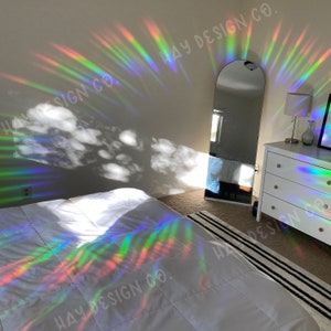 Puede incluir: Un interior de dormitorio con una cama blanca, un espejo de cuerpo entero y una c&oacute;moda. La habitaci&oacute;n est&aacute; decorada con un prisma de arco&iacute;ris que proyecta patrones de luz coloridos en las paredes y los muebles. El prisma est&aacute; etiquetado con "HAY DESIGN CO."