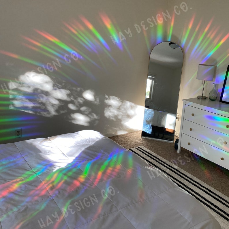 Puede incluir: Un interior de dormitorio con una cama blanca, un espejo de cuerpo entero y una c&oacute;moda. La habitaci&oacute;n est&aacute; decorada con un prisma de arco&iacute;ris que proyecta patrones de luz coloridos en las paredes y los muebles. El prisma est&aacute; etiquetado con "HAY DESIGN CO."