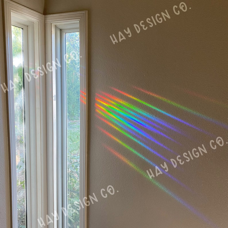 Puede incluir: Un prisma de arco&iacute;ris colgado en una ventana proyecta rayos de luz coloridos en una pared con el texto "HAY DESIGN CO." repetido en diferentes posiciones.