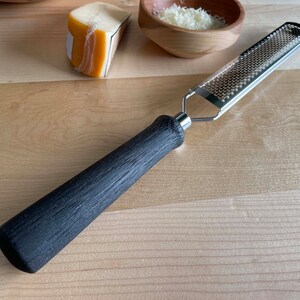 May include: A stainless steel grater with a black textured handle. The grater is designed for fine grating and is ideal for cheese or other ingredients. A wedge of cheese and a bowl of grated cheese are also visible.