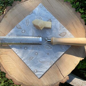May include: A stainless steel grater with a wooden handle and a piece of cheese on a patterned napkin. The grater is designed for fine grating and is ideal for cheese or other ingredients. The napkin has a floral design.