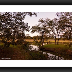 May include: A scenic view of a creek winding through a field with trees lining the banks. The trees are mostly bare, with some leaves still clinging to the branches. The sky is a soft blue with a hint of pink in the clouds.