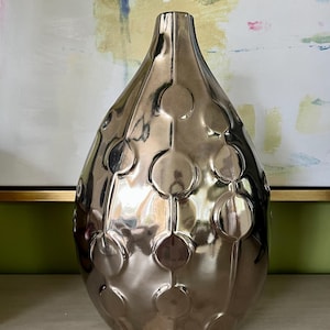 May include: A teardrop-shaped vase with a reflective metallic bronze finish. The surface features a repeating pattern of raised circular discs connected by vertical lines, creating a textured, geometric design across the entire exterior.
