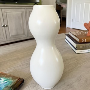 May include: A smooth, off-white ceramic vase with a distinct hourglass or gourd-like silhouette, featuring a narrow, cinched middle section that flares out into rounded top and bottom bulbs, finished with a simple, circular opening at the top.