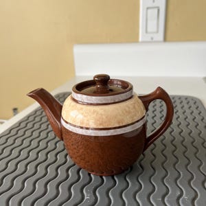 May include: A brown and beige ceramic teapot with a lid and handle. The teapot has a textured lower half and a smooth, striped upper half. The spout and handle are a darker brown. A classic kitchenware item.