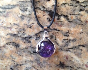 May include: A silver pendant necklace with a purple amethyst gemstone held in a pair of silver hands. The necklace is on a black cord.