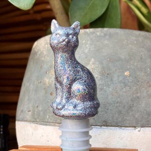 May include: A silver glitter cat bottle stopper. The cat is in a seated position with its ears perked up. The stopper is made of a silver glitter material with iridescent highlights. The stopper is attached to a white and clear plastic base.