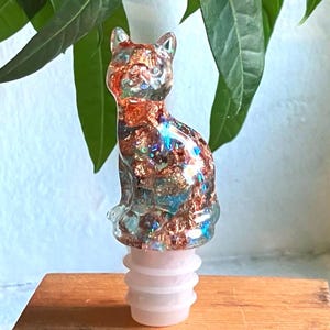 Puede incluir: Un tapón de botella de vidrio con forma de gato con purpurina de cobre y azul incrustada en el vidrio. El tapón es blanco y tiene un diseño roscado.