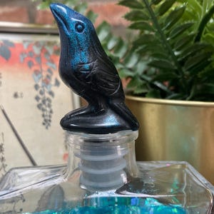 May include: A decorative bottle stopper featuring a black raven with iridescent blue highlights. The raven is perched atop a clear glass bottle with a clear plastic stopper. The bottle contains blue glitter.