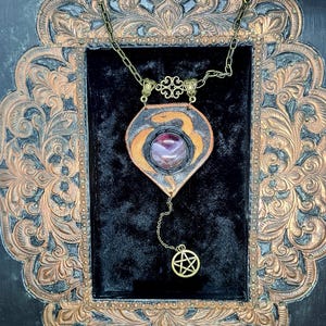 May include: A handcrafted pendant necklace featuring a central purple gemstone set in a leather and metal pendant. The pendant hangs from a chain with decorative elements and a dangling pentagram charm. The necklace is displayed on a black velvet background.