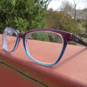 Puede incluir: Gafas con montura degradada, pasando de rojo oscuro a azul claro. Las patillas son negras y las lentes transparentes. Las gafas est&aacute;n apoyadas en una superficie.
