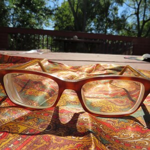 May include: Brown rectangular eyeglasses with clear lenses rest on a colorful paisley patterned fabric. The frames have a tortoiseshell design. The background includes a wooden deck and trees, suggesting an outdoor setting.