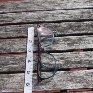 Puede incluir: Gafas con montura burdeos oscuro y azul, de aproximadamente 14 cm de largo, colocadas sobre una superficie de madera envejecida. Una cinta m&eacute;trica blanca se coloca junto a las gafas como referencia de tama&ntilde;o. Las lentes son transparentes.