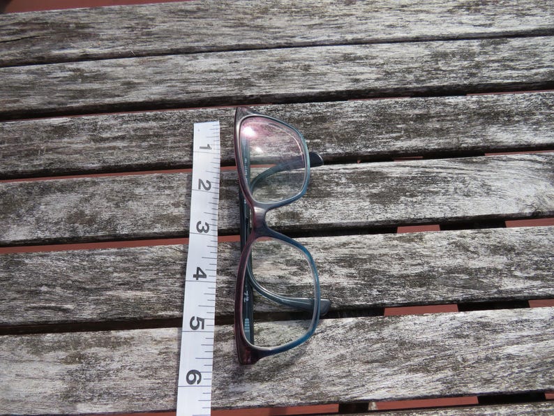 Puede incluir: Gafas con montura burdeos oscuro y azul, de aproximadamente 14 cm de largo, colocadas sobre una superficie de madera envejecida. Una cinta m&eacute;trica blanca se coloca junto a las gafas como referencia de tama&ntilde;o. Las lentes son transparentes.