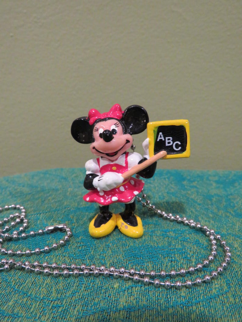 Peut inclure: Un collier pendentif Minnie Mouse. Le pendentif repr&eacute;sente Minnie Mouse tenant un tableau avec "ABC" dessus. Minnie porte une robe &agrave; pois rouges et blancs, un n&oelig;ud rose et des chaussures jaunes. Le collier a une cha&icirc;ne &agrave; billes argent&eacute;e.