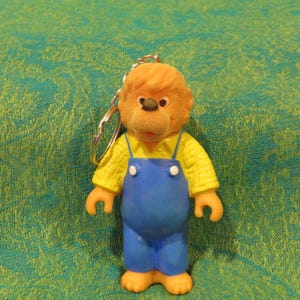 May include: A vintage keychain featuring a cartoon bear character. The bear has a fuzzy orange head, a yellow shirt, and blue overalls. The keychain is made of plastic and metal, with a silver-colored chain.