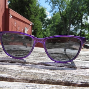 May include: A pair of purple sunglasses with a translucent frame rests on a weathered wooden surface. The lenses are dark, and the sunglasses are positioned to catch the sunlight. The background includes a red fence and green trees.