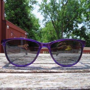 May include: A pair of purple sunglasses with a rectangular frame and dark lenses. The sunglasses are resting on a weathered wooden surface, reflecting the surrounding environment. The image is taken outdoors on a sunny day.