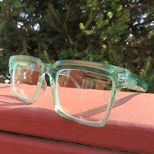 May include: A pair of clear-lensed eyeglasses with a light green, square-shaped frame. The glasses are resting on a red surface, with a backdrop of green foliage. The frames have a textured design on the temples.