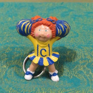 May include: A Cabbage Patch Kids cheerleader keychain. The figurine has red hair, freckles, and is wearing a yellow and blue uniform with the letter "C". The cheerleader is holding up blue and yellow pom-poms.