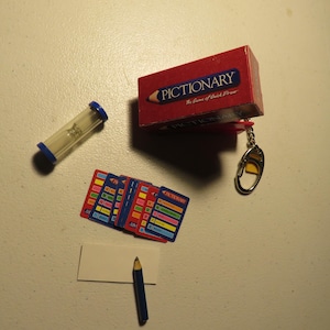 May include: A red and blue Pictionary game box with a pencil and a keychain. The box is open and shows the game's name and the words "The Game of Quick Draw". There is also a blue hourglass, a pencil, and a stack of cards.