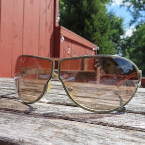 May include: Vintage aviator sunglasses with a gold-tone metal frame and gradient brown lenses. The sunglasses are resting on a weathered wooden surface, with a red wooden structure in the background. The lenses reflect the sky and surrounding trees.