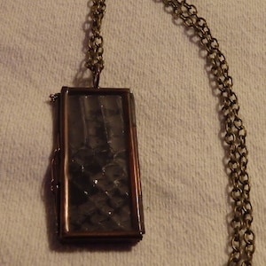 May include: A gold chain necklace with a rectangular locket pendant. The locket has a clear glass front and a dark patterned design inside.
