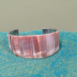 May include: A cuff bracelet featuring a series of vertical, rectangular shell segments in shades of pale pink and deep reddish-brown. The segments display natural linear patterns and are set against a metallic silver-toned interior band.