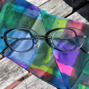 May include: Oval-framed eyeglasses with a silver-colored metal frame rest on a colorful, abstract patterned fabric. The fabric features a mix of blues, greens, purples, and pinks. The glasses have clear lenses and black earpieces. The text on the glasses reads "VFPAGE 47018 289P-121 137".