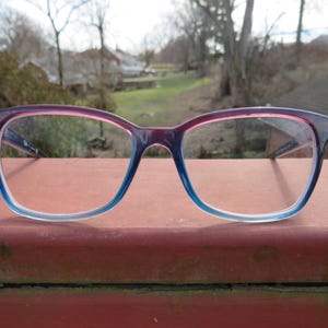 Puede incluir: Un par de gafas con montura degradada. La parte superior de la montura es de color rosa intenso que se desvanece en azul claro en la parte inferior. Las lentes son transparentes y las gafas est&aacute;n sobre un fondo exterior borroso.