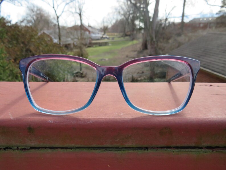 Puede incluir: Un par de gafas con montura degradada. La parte superior de la montura es de color rosa intenso que se desvanece en azul claro en la parte inferior. Las lentes son transparentes y las gafas est&aacute;n sobre un fondo exterior borroso.
