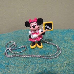 Peut inclure: Figurine de Minnie Mouse v&ecirc;tue d'une robe rose &agrave; pois et tenant un petit tableau avec "ABC" &eacute;crit dessus. La figurine a un collier en cha&icirc;ne &agrave; billes argent&eacute;es. Le fond est un tissu turquoise.