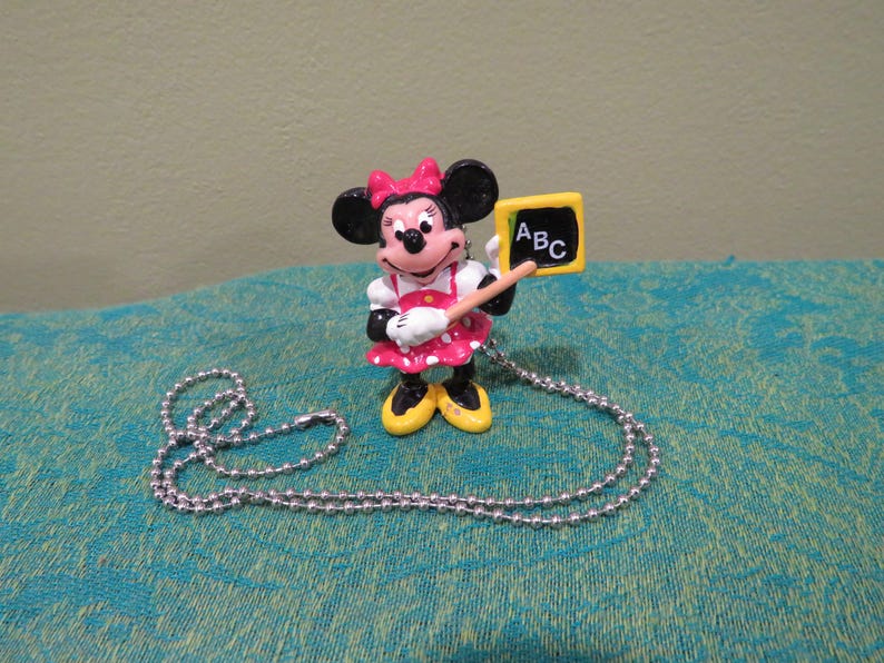 Peut inclure: Figurine de Minnie Mouse v&ecirc;tue d'une robe rose &agrave; pois et tenant un petit tableau avec "ABC" &eacute;crit dessus. La figurine a un collier en cha&icirc;ne &agrave; billes argent&eacute;es. Le fond est un tissu turquoise.