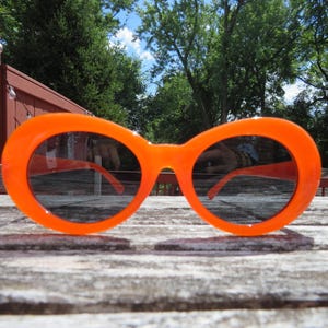 May include: Bright orange sunglasses with large, round frames and dark lenses. The sunglasses are set against a wooden table, with a background of trees and a blue sky. The frames are a solid, vibrant color.