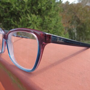 Puede incluir: Gafas rectangulares con montura burdeos y azul claro. Las patillas son de color azul marino oscuro. Las lentes son transparentes. Las gafas est&aacute;n sobre una superficie.