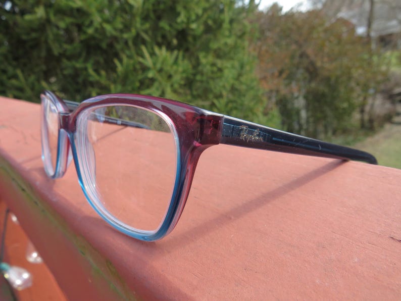 Puede incluir: Gafas rectangulares con montura burdeos y azul claro. Las patillas son de color azul marino oscuro. Las lentes son transparentes. Las gafas est&aacute;n sobre una superficie.