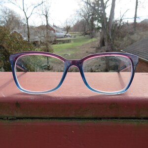 Puede incluir: Gafas con montura degradada, que va del rosa al azul. Las gafas est&aacute;n sobre una superficie roja, con un fondo exterior borroso de &aacute;rboles y edificios.