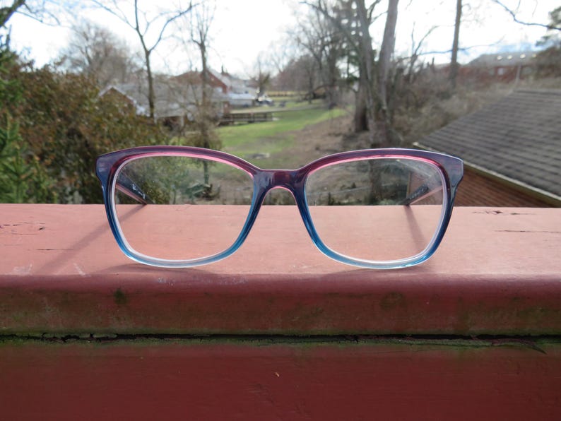 Puede incluir: Gafas con montura degradada, que va del rosa al azul. Las gafas est&aacute;n sobre una superficie roja, con un fondo exterior borroso de &aacute;rboles y edificios.