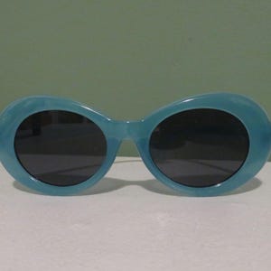 May include: Oval-shaped sunglasses in a light blue hue. The frames have a marbled appearance, and the lenses are dark. The sunglasses are set against a white surface with a green background.