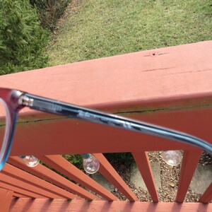 Puede incluir: Primer plano de gafas con montura roja y azul. Las gafas est&aacute;n sobre una superficie de madera roja, con un fondo verde de hierba y &aacute;rboles. Las gafas tienen forma rectangular.