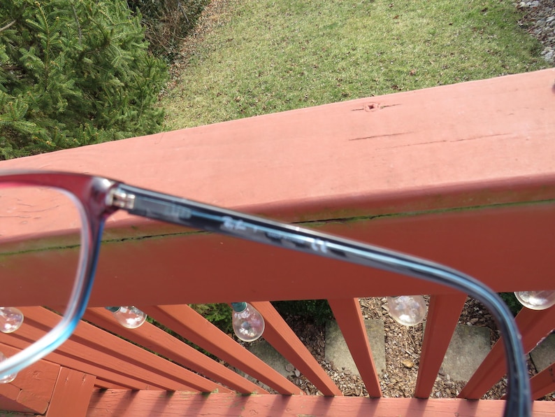 Puede incluir: Primer plano de gafas con montura roja y azul. Las gafas est&aacute;n sobre una superficie de madera roja, con un fondo verde de hierba y &aacute;rboles. Las gafas tienen forma rectangular.